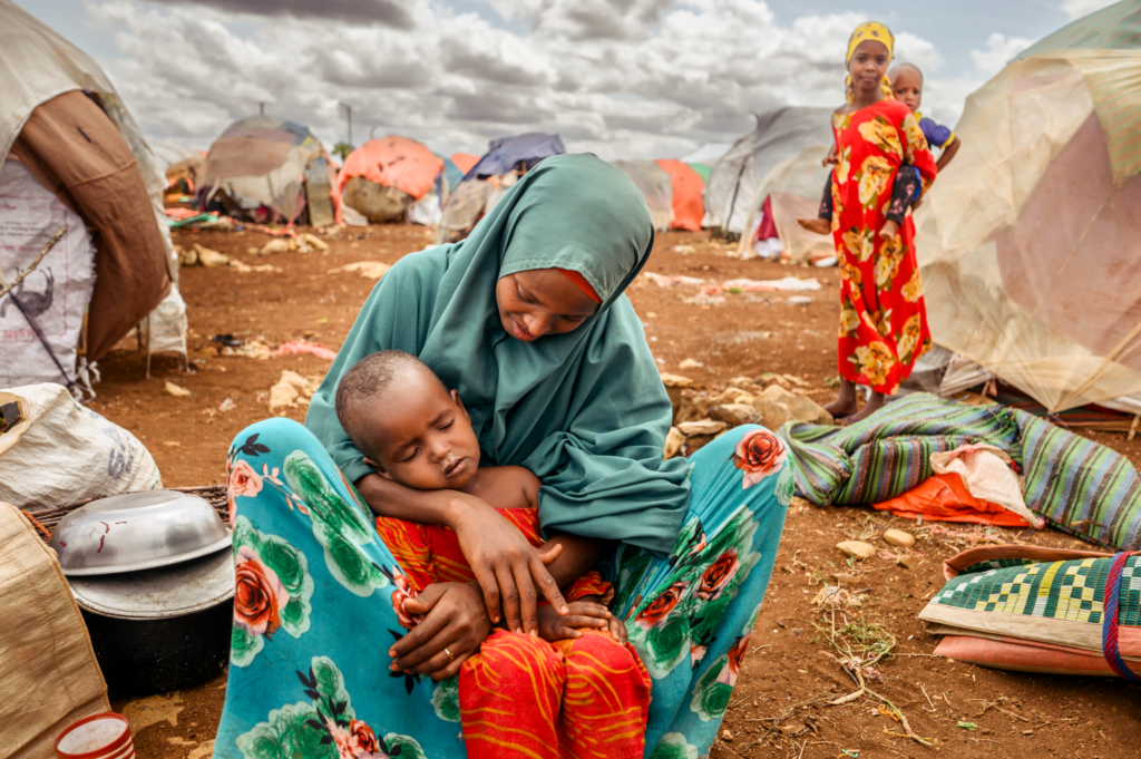 Somalilainen äiti pitää tuskallisen näköisenä lapsensa sylissä. Taustalla näkyy pakolaisleiri.