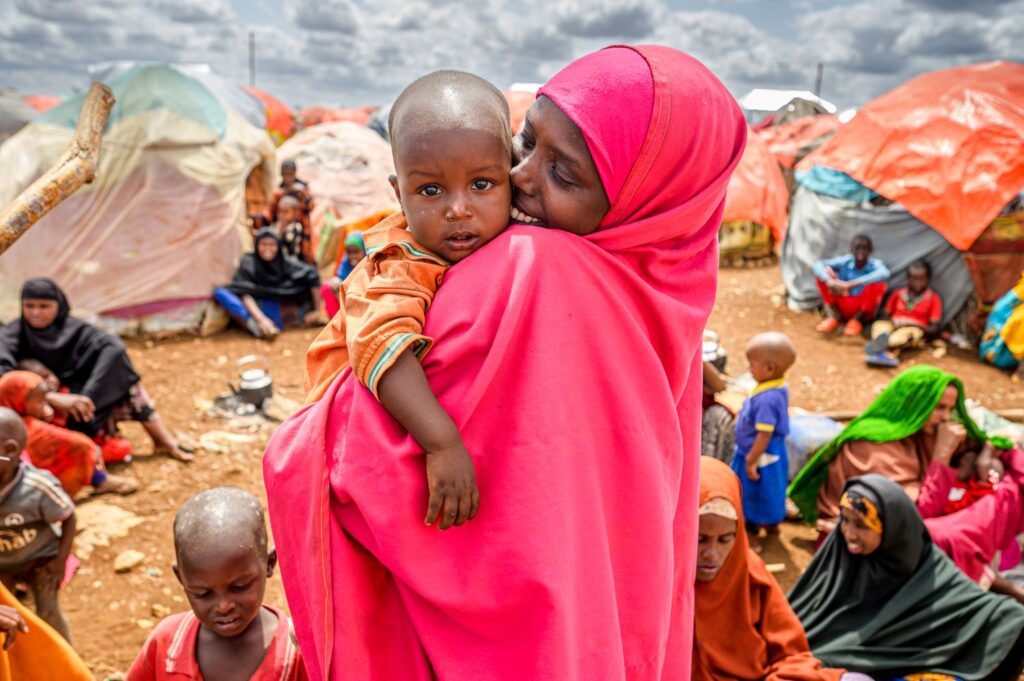 Somalilainen äiti halailee taaperonsa hynyillen. Taustalla näkyy pakolaisleiri.