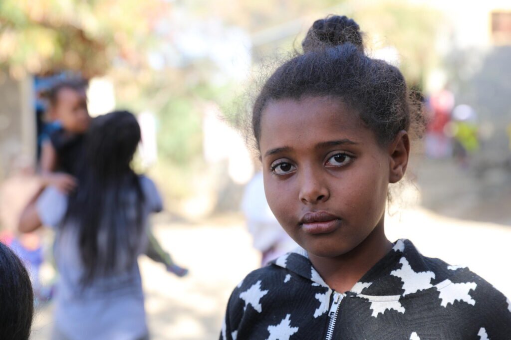 Etiopialainen tyttö katsoo kameraan. Taustalla näkyy nainen kantamassa lasta sylissään.