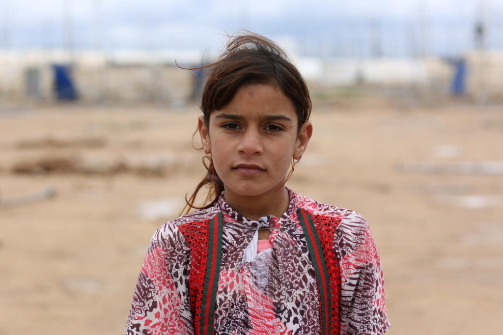 Irakilainen tyttö katsoo vakavana kameraan. Taustalla näkyy pakolaisleiri.