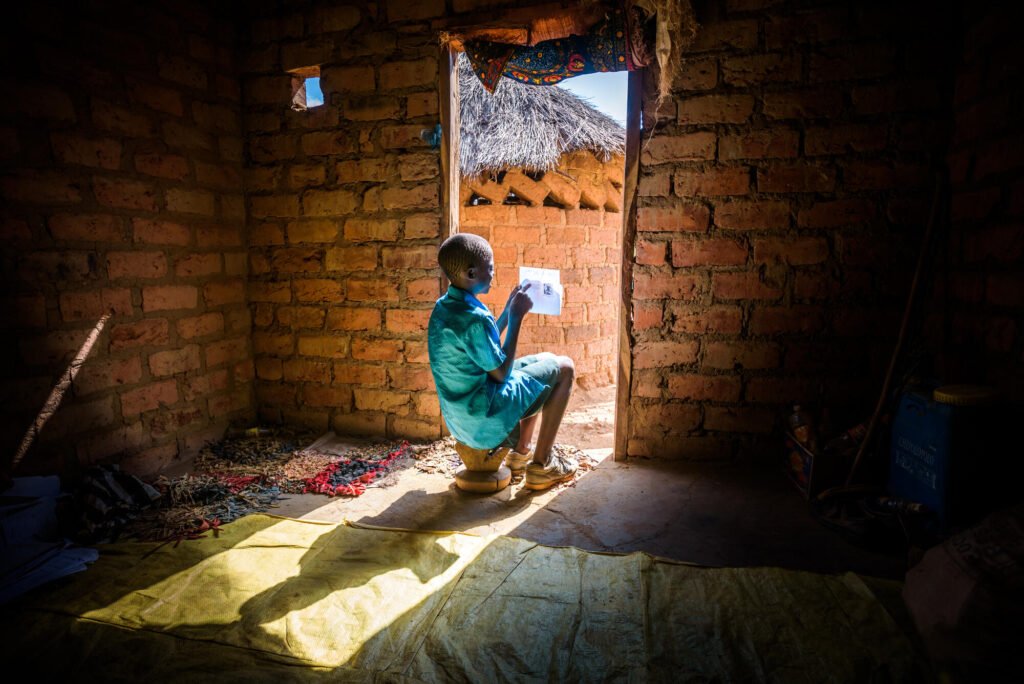 Sambialainen kummilapsi istuu kotinsa ovensuussa ja pitelee kädessään kirjettä. Ulkoota paistaa valoa sisälle ja lapsi istuu auringossa. Kuva on otettu takaapäin.