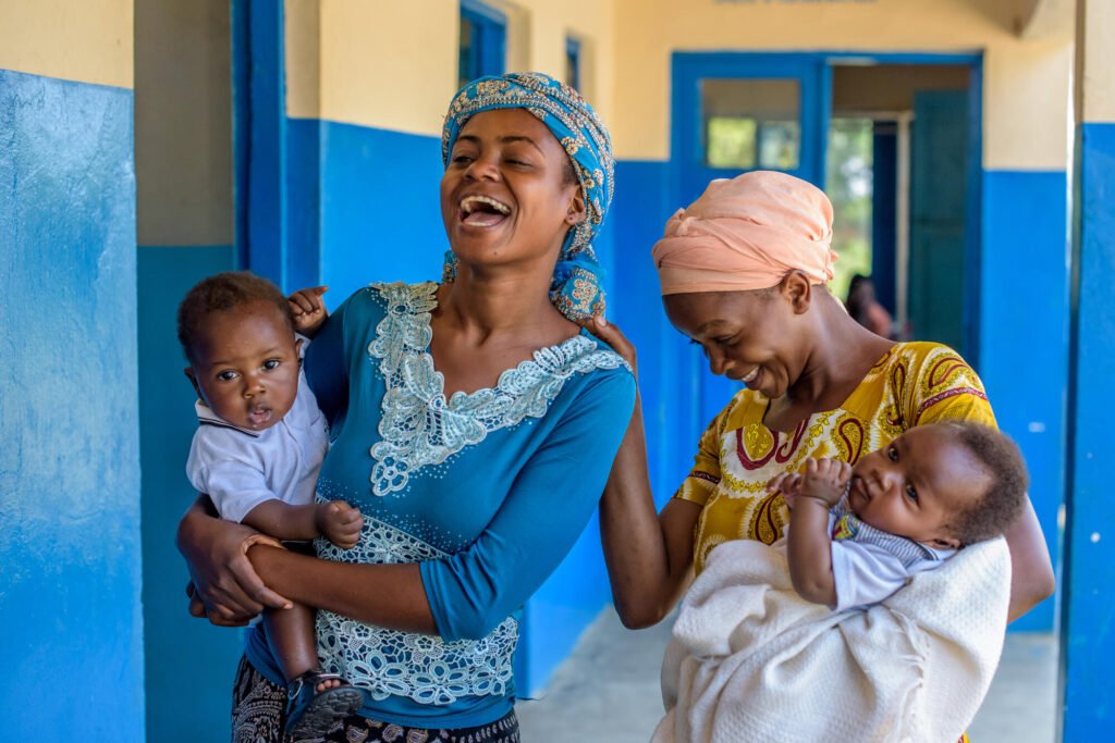 Kaksi kongolaista äitiä nauravat yhdessä, terveysasemalla, taaperot sylissään.
