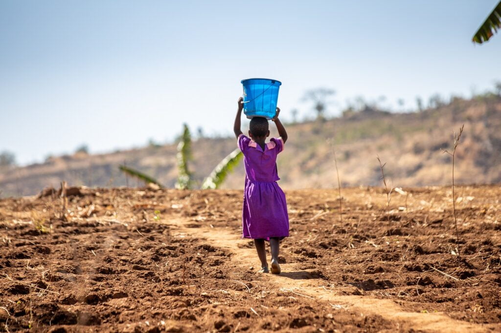 Kenilalainen pikkutyttö kantaa pään päällä vesiämpärin ja kävelee kuivan maaston läpi.