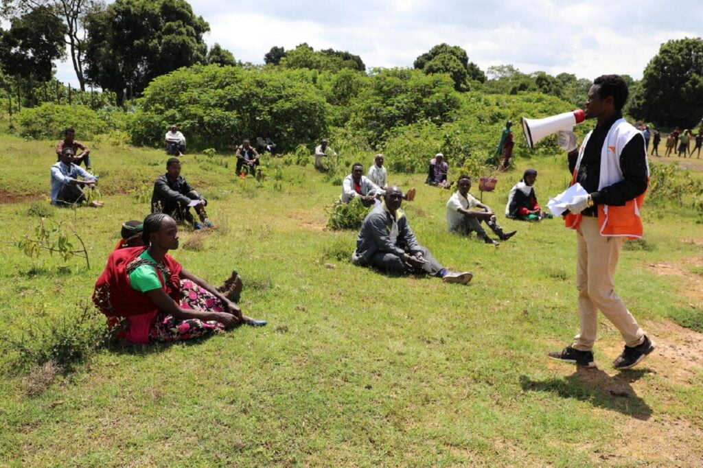 World Visionin paikallinen työntekijä puhumassa megafoniin. Edessä turvaväleillä istuu ihmisiä ruoholla kuuntelemassa.