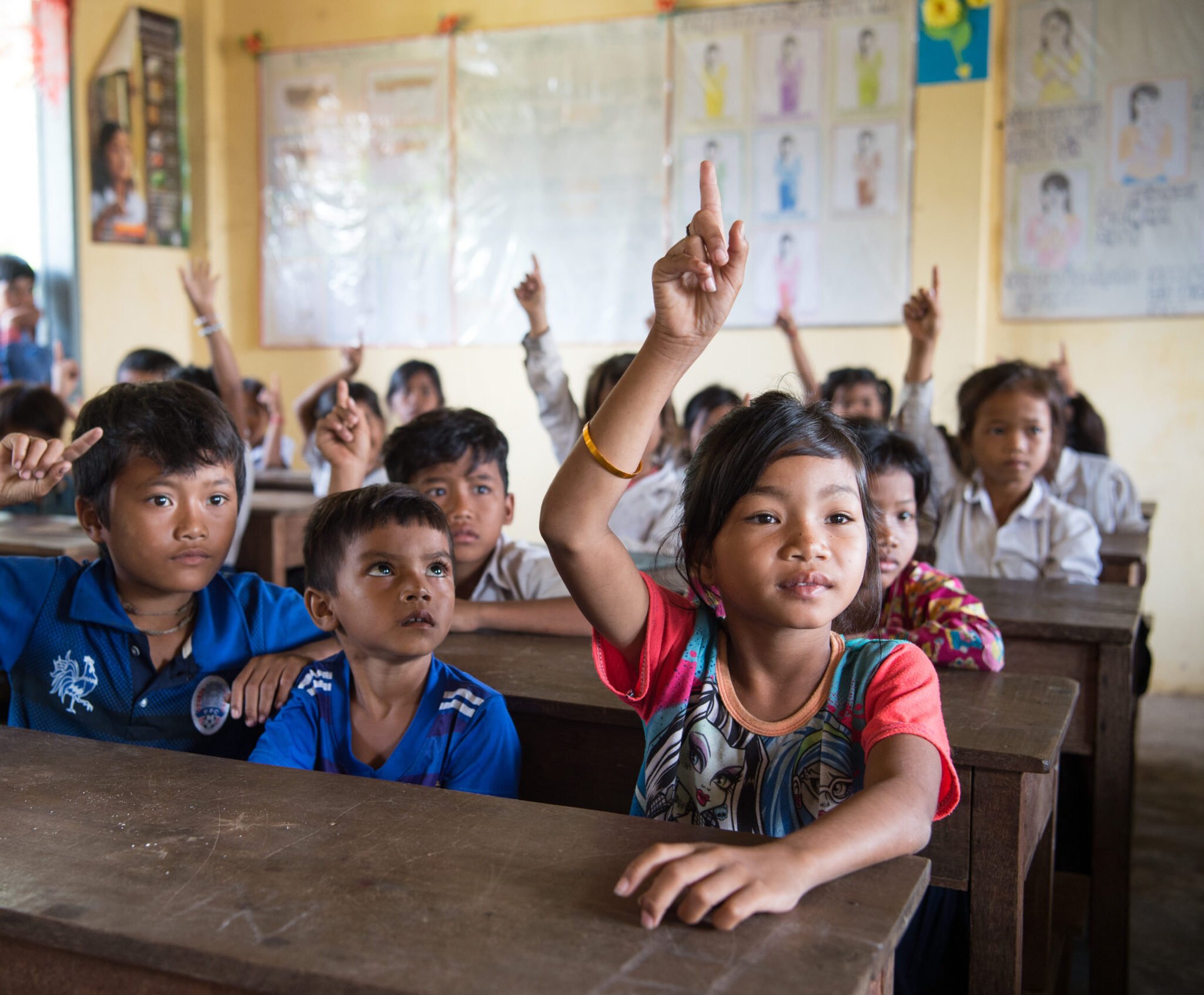 Kambodzalainen koulutyttö viittaa luokkahuoneessa. Ympärillä näkyy muita koululaisia.