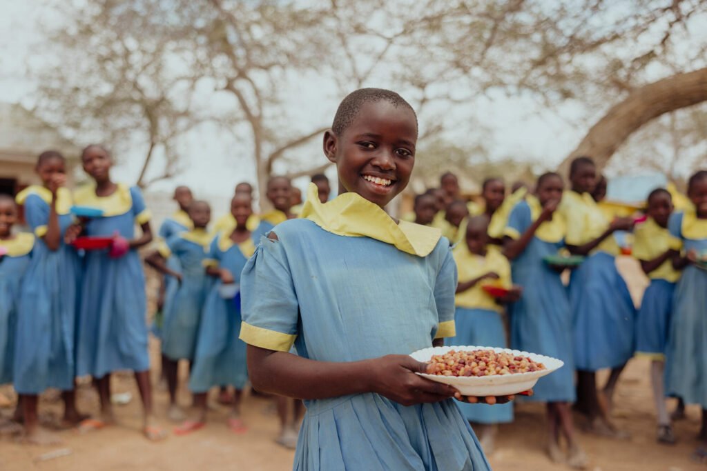 Kenialainen koulutyttö nauttii kouluruuasta ja hymyilee kameralle. Taustalla näkyy muita koululaisia.