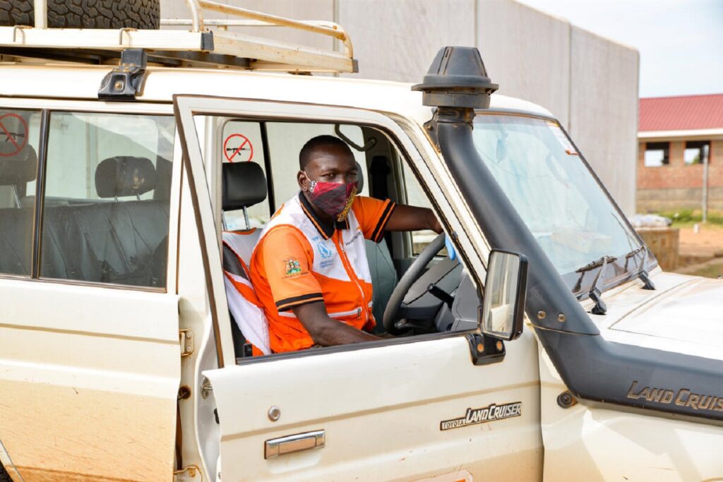 World Visionin paikallinen työntekijä istuu auton ratissa, katsomassa kameraan kasvomaski päällä.