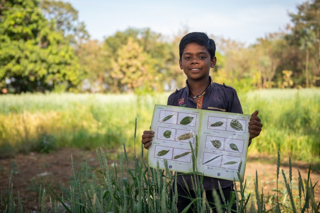 Intialainen poika seisoo pellolla ja esittelee tehtäväkirjaa, jossa on puiden lehtiä.