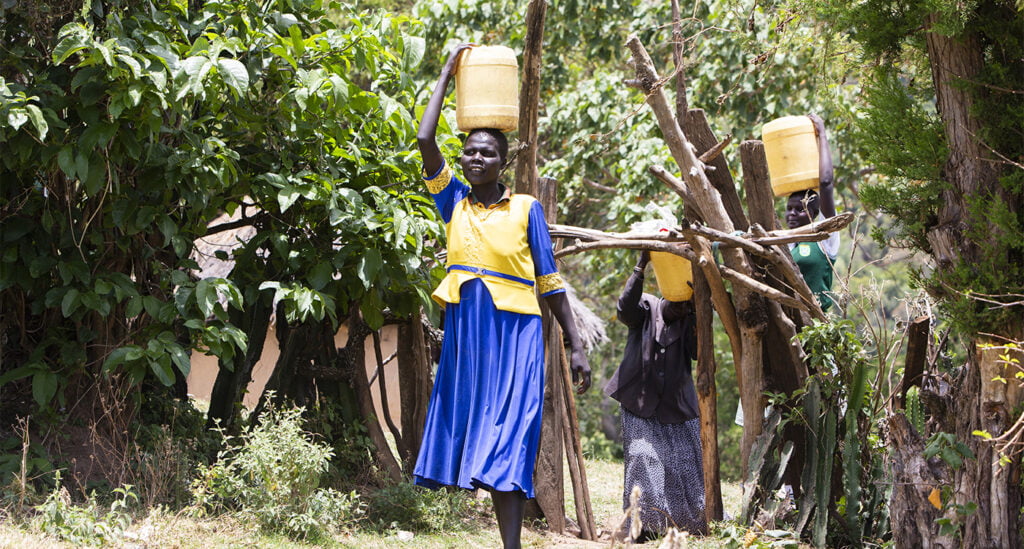 Äiti Domtilla Loskolian kantaa vettä kanisterissa päällään. Hän on pukeutunut siniseen ja keltaiseen mekkoon. Taustalla näkyy isoäiti Cheptoi sekä tytär Joylene, jotka myös kantavat vettä.