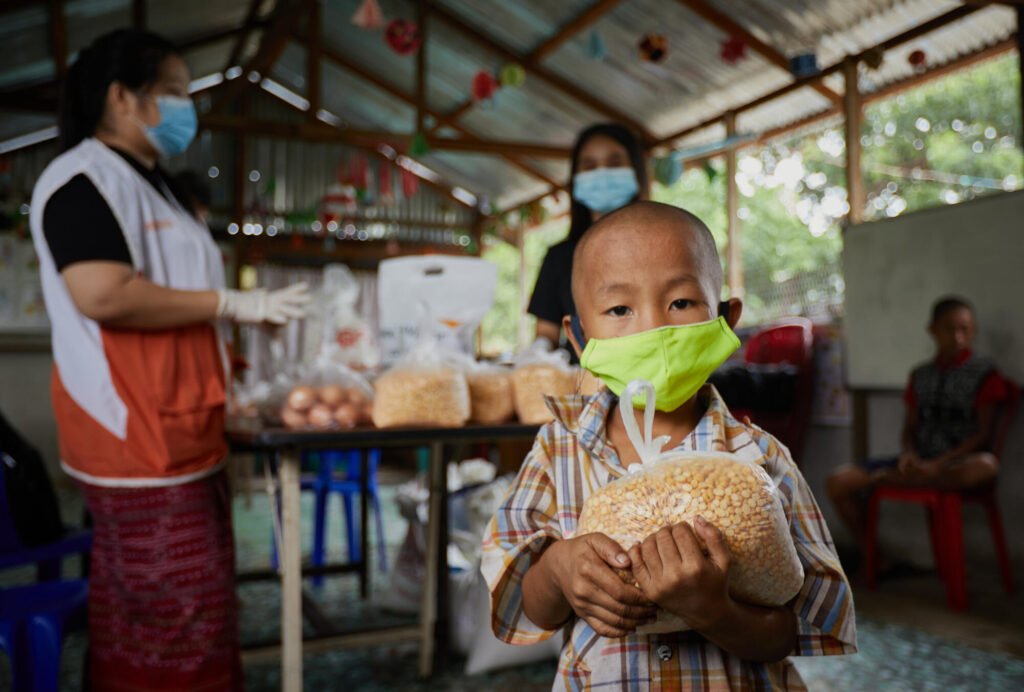 Myanmarilainen poika pitää ruokapussia sylissä ja katsoo kameraan, kasvomaski päällä.