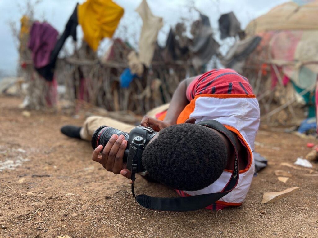 World Visionin liiviin pukeutunut valokuvaaja makaa maassa ottamassa kuvaa.