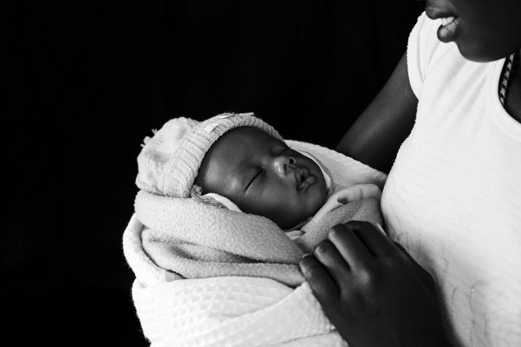 Mustavalkoinen lähikuva nukkuvasta, vastasyntyneestä kenialaisesta vauvasta äitinsä sylissä.