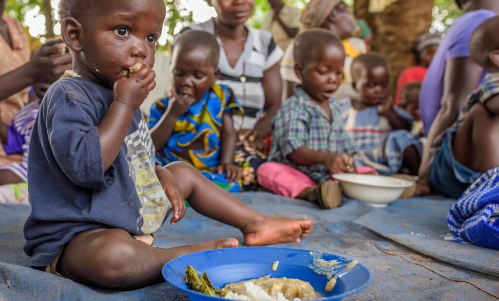 Ugandalainen taapero istuu viltillä ja syö ruokaa lautaselta. Taustalla näkyy muita taaperoita ja heidän äitejään.