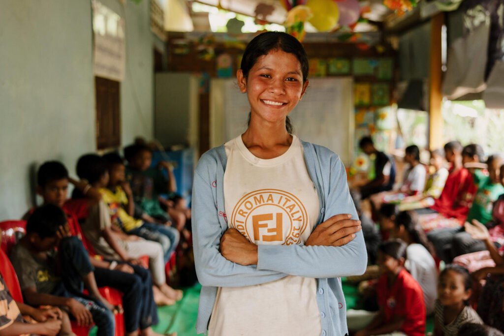 Kambodzalainen tyttö hymyilee kameralle, luokkatovereita takanaan.