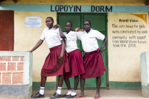 Kolme kenialaista koulutyttöä poseeraavat itsevarmasti turvatalon edessä. Talon seinällä on World Visionin logo sekä teksti englanniksi, joka kertoo, että tämä tyttöjen turvatalo on rakennuttanut Sookin yhteisö World Vision Kenian tuella vuonna 2013.