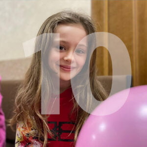 Ukrainalainen tyttö hymyilee kameralle, ilmapallo vieressään. Kuvan päällä on numero 10.