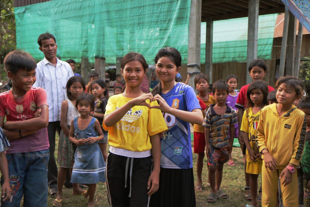 Kambozalaisia lapsia seisovat yhdessä ulkona. Keskellä on kaksi tyttöä, jotka seisovat vierekkäin ja muodostavat käsillään sydän yhdessä.