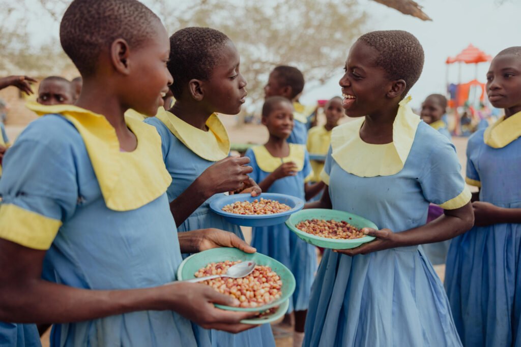 Kenialaiset koulutytöt nauttivat kouluruuasta ja nauravat yhdessä. Taustalla näkyy muitakin koululaisia syömässä.