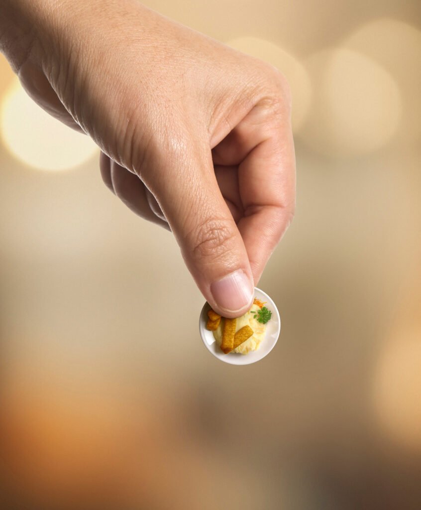 Käsi pitää sormien välissä kiinni kahden euron kolikon, mutta kun katsoo tarkemmin, kolikko osoittautuu lautaseksi, jossa on perunamuussia sekä kalapuikkoja numero 2-muodossa.